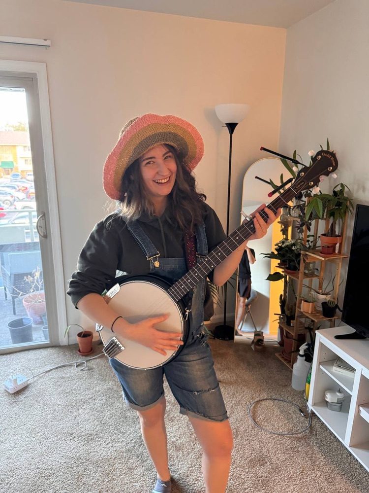 Buse smiling and holding a banjo in a living room, wearing a striped knit hat.