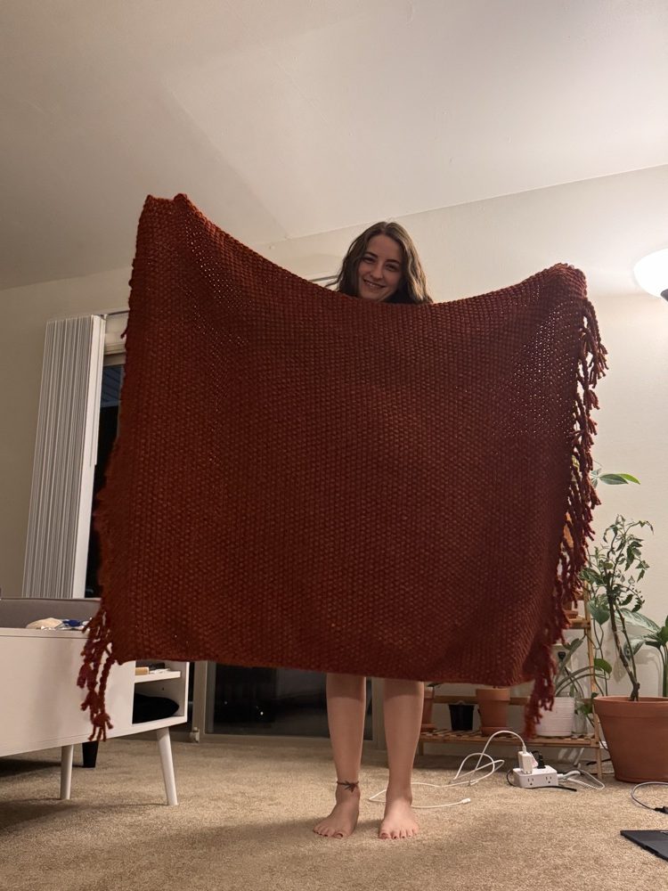 Buse holding up a large hand-knit rust-colored blanket with fringe.